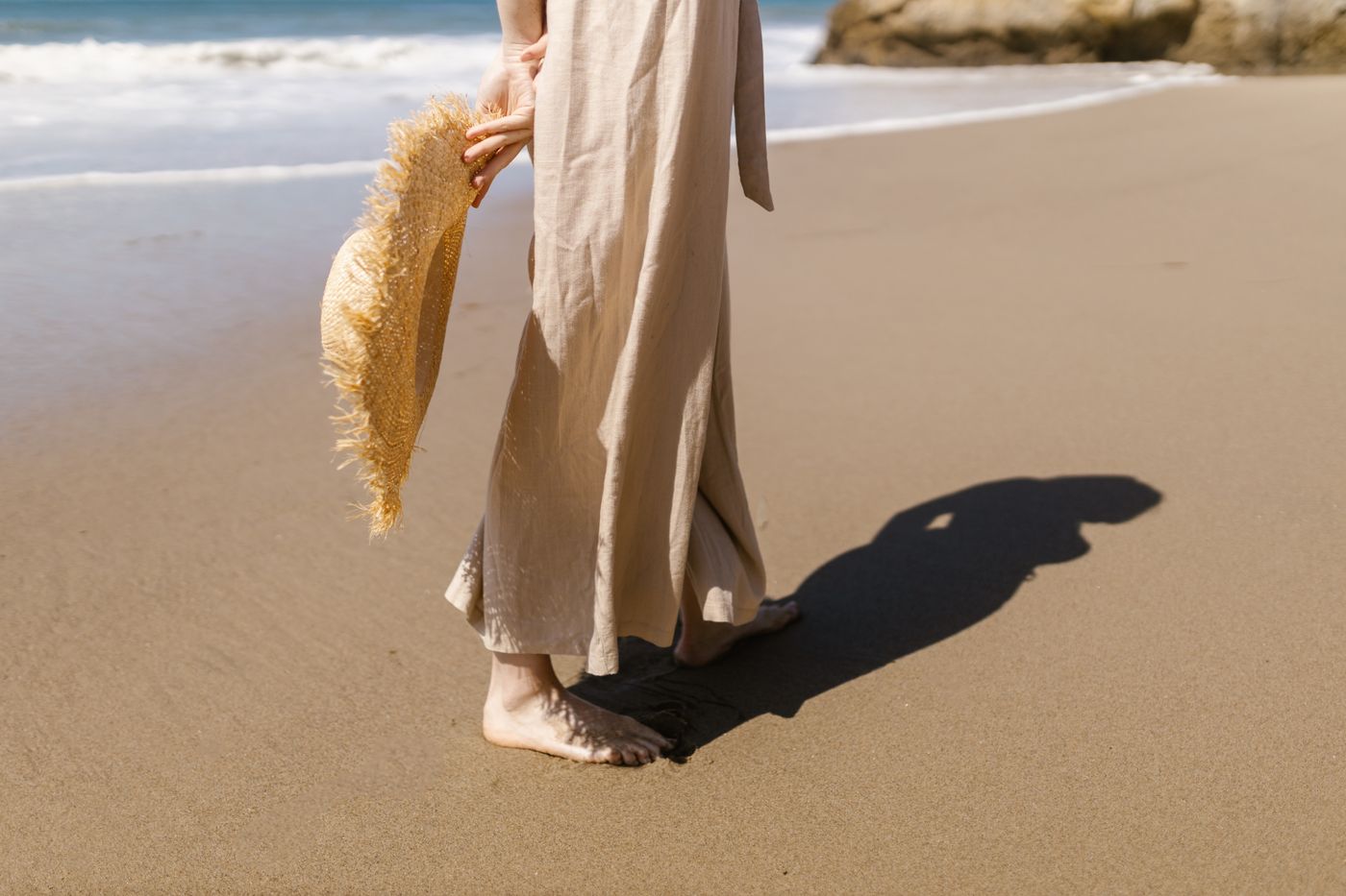 Woman wearing loose beige trousers while walking barefoot on a beach.