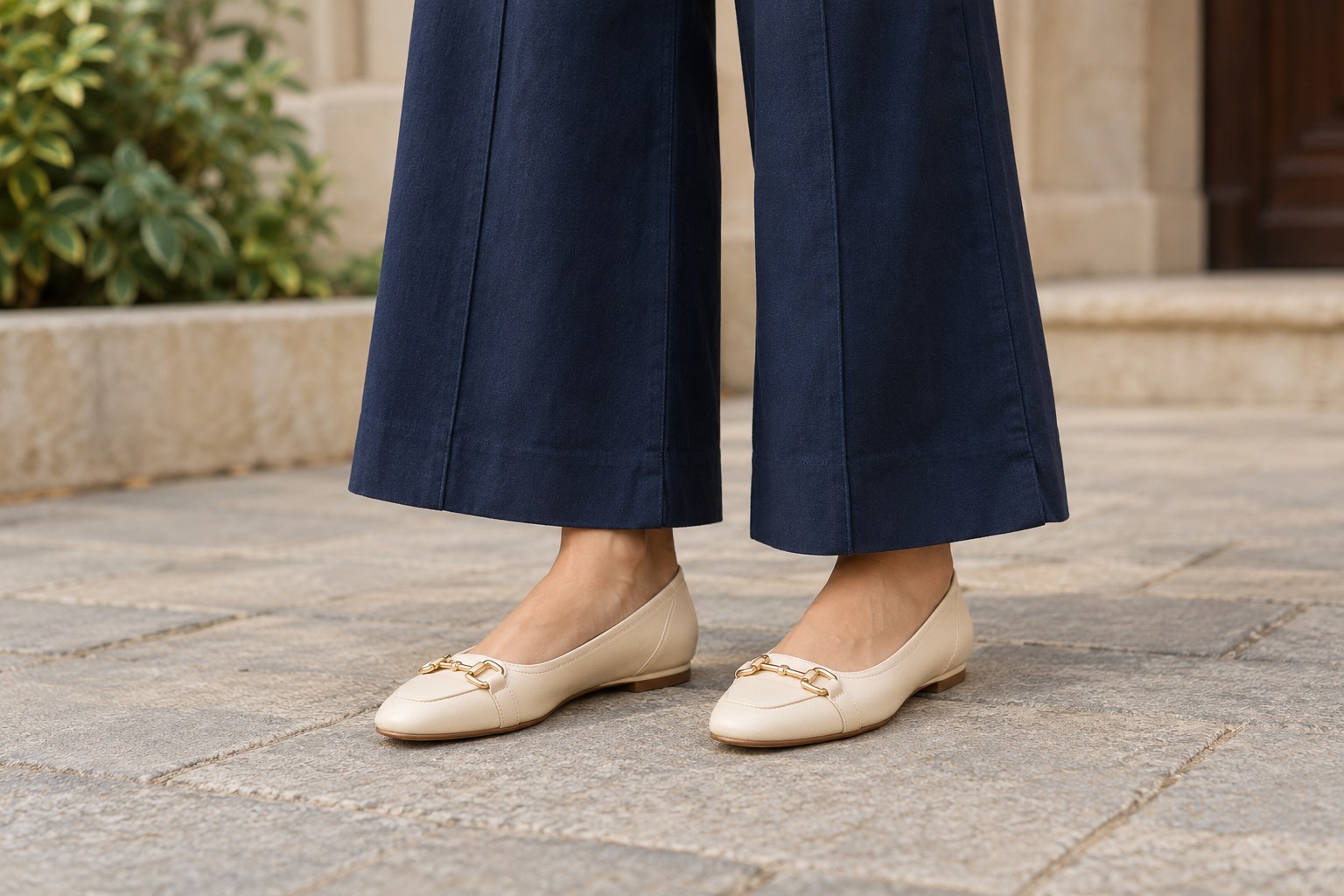 Lower half of adult Indian woman wearing wide-leg navy trousers ending above cream leather flat shoes, full shoes and trouser hem visible.