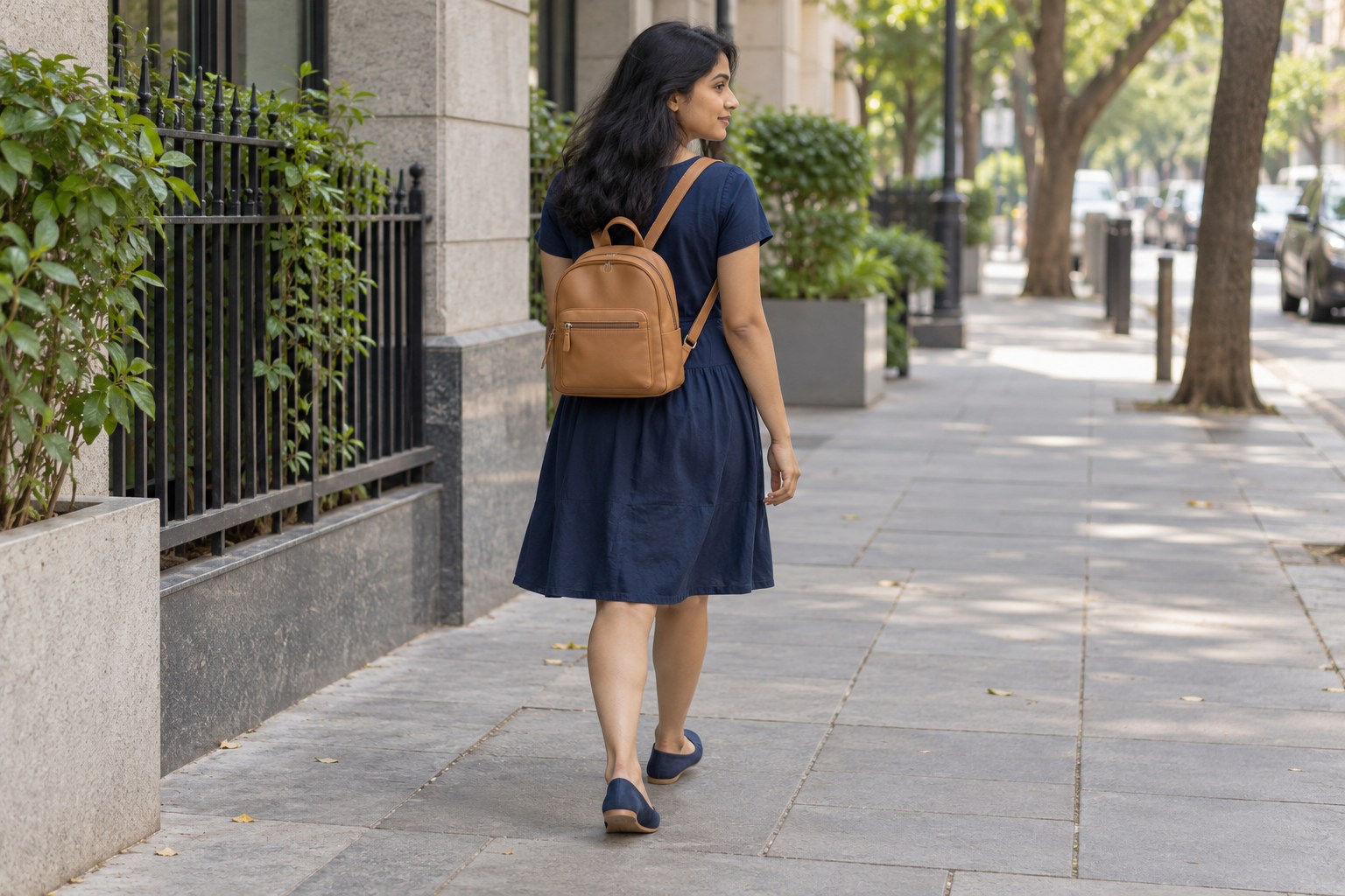 Woman wearing a navy knee-length dress and tan backpack while walking on a city pavement.
